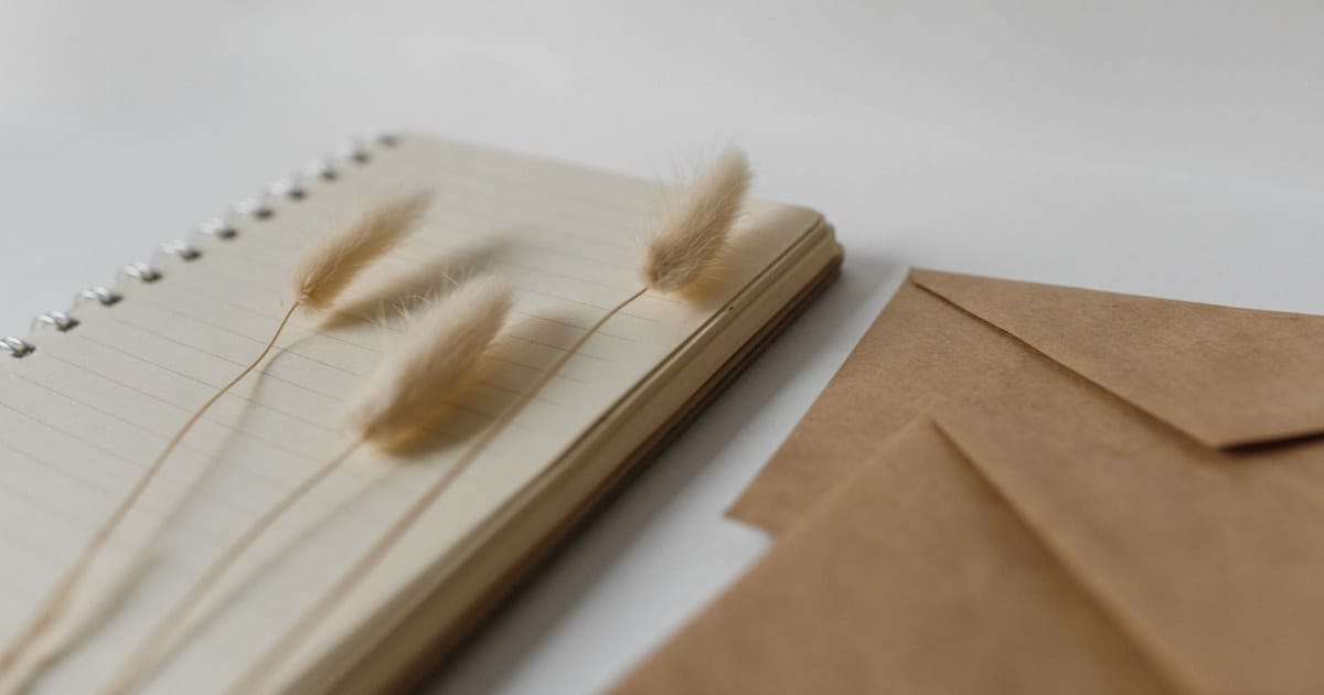 Un quaderno aperto con dei fiori secchi appoggiati sopra e due buste di carta accanto, su un piano chiaro