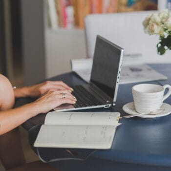 Donna che scrive appunti a mano su un quaderno accanto al computer, con una tazza di caffè sul tavolo